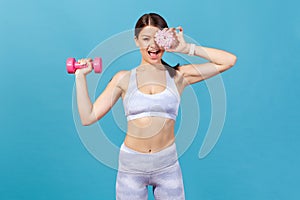 Cheerful athletic woman in white tights and top having fun, closing eye with donut, holding dumbbell in hand, burning extra