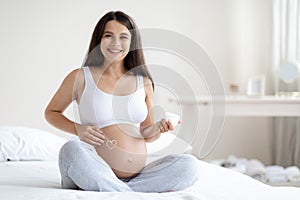 Cheerful attractive pregnant woman massaging her big belly, using cream