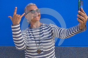 Cheerful attractive senior woman with white hair smiling happy making a selfie using smart phone standing - blue background - tech