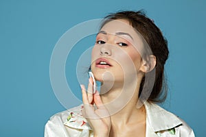 Cheerful attractive teen girl clean her face with cotton disk, isolated over blue background. Portrait of young beautiful woman