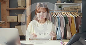 Cheerful attractive young Asian lady with casual smile show open sign in shop reopen from coronavirus pandemic at her cloth shop.
