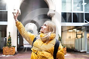Cheerful attractive young blonde woman wearing yellow jacket with backpack walking in the city