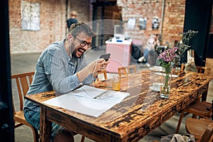 Cheerful attractive young man with glasses and earphones in cafe, using smarphone laughing, video calling, audio calling photo