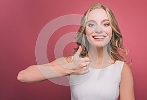 Cheerful and attractive young person is holding her thumb up and smiling. She is very cheerful. Isolated on pink