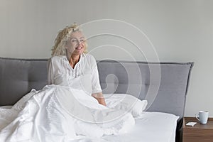 Cheerful awaking pretty mature woman sitting in comfortable soft bed