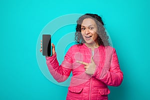 Cheerful awesome curly haired charming woman in bright pink jacket looks at camera, pointing her finger on a blank screen of a