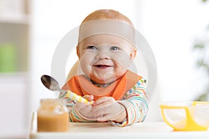 Cheerful baby child eats food itself with spoon. Portrait of happy kid boy in high-chair.