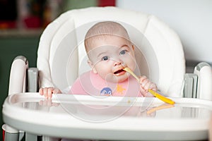 Cheerful baby child eats food itself with spoon. Portrait of happy kid girl in high chair.