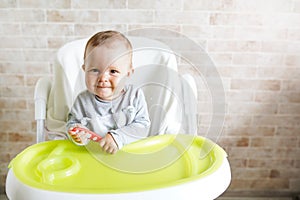 Cheerful baby child eats food itself with spoon. Portrait of happy kid in high chair in sunny kitchen. background with copy space