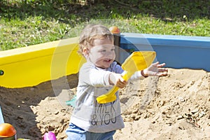 Cheerful baby girl boy playing with a shovel in the sandbox. Laughing face smiling child kid in a gray sweater walks on the