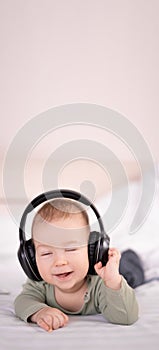 Cheerful baby listening to music in headphones lying on white bed, toddler 3 months old little dj, happy childhood