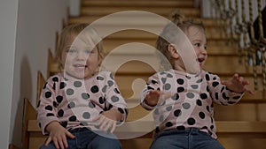 Cheerful baby twins sitting on stairs at home laughing gesturing in slow motion. Portrait of joyful positive happy