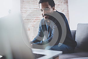 Cheerful bearded African guy working on laptop while sitting sofa at his modern office place.Concept of young people