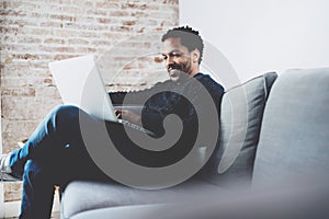 Cheerful bearded African man working on laptop while sitting sofa at his modern office place.Concept of young people