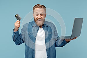 Cheerful bearded hipster blonde young man in jeans shirt holding notebook and credit card over blue background.