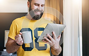 Cheerful bearded man stands and using tablet computer. Guy laughs looking screen of digital tablet while drinking coffee