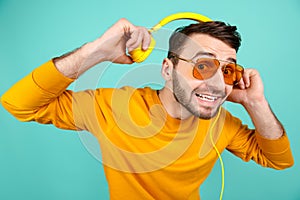 Cheerful bearded young man wearing yellow sunglasses listening to music with yellow headphones on cyan background.