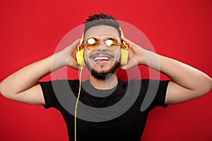 Cheerful bearded young man wearing yellow sunglasses listening to music with yellow headphones on red background.