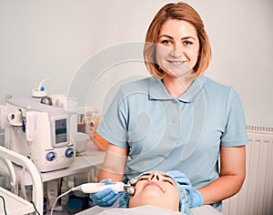 Cheerful beautician massaging woman face with facial roller.