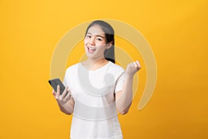 Cheerful beautiful Asian woman holding smartphone on light yellow background.
