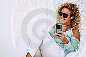 Cheerful beautiful curly young adult woman use mobile phone outdoor sit down on a white garden elegant chair -smile and enjoy the