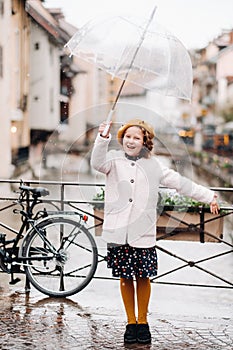 Cheerful beautiful girl in a coat with a transparent umbrella in Annecy. France. The girl Cheerfully raises an umbrella in the