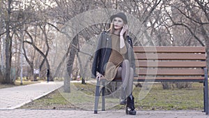 Cheerful beautiful girl in hat sitting on bench in sunny autumn park. Action. Beautiful young woman in hat sitting on