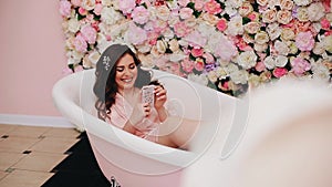 Cheerful beautiful girl lying in a white tub with a smartphone in hand.
