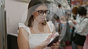 Cheerful, beautiful hispanic woman wearing glasses, happily using smartphone while standing alone on the empty subway - a portrait