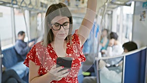 Cheerful, beautiful hispanic woman wearing glasses, happily using smartphone while standing alone on the empty subway - a portrait