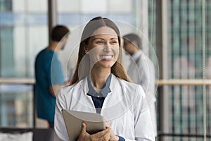 Cheerful beautiful practitioner woman holding tablet, standing in hospital hall