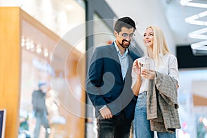 Cheerful beautiful young couple using mobile phone standing in hall of shopping mall. Bearded man in glasses and blonde