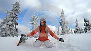 Cheerful beautiful young girl snowboarder runs in the snow among snowy pines. Falls to his knees, throws snow flakes