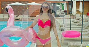 Cheerful beautiful young woman walking by the pool holding sprinkled pink inflatable flamingo float in summer. Pool