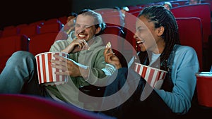 Cheerful best friends laughing, having popcorn while watching movie together, sitting in cinema auditorium