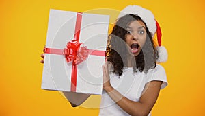 Cheerful biracial girl in santa claus hat saying Wow and showing huge giftbox