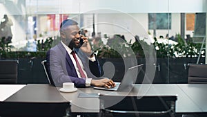 Cheerful black businessman working on laptop, having phone conversation