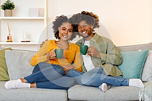 Cheerful Black Couple Hugging Holding Coffee Cups Sitting At Home