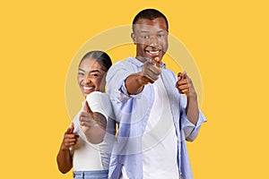 Cheerful black couple pointing at viewer, engaging smiles, yellow