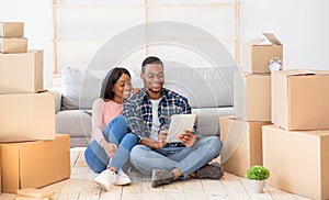 Cheerful black couple with touch pad to search for household goods for their new home online, using tablet on moving day