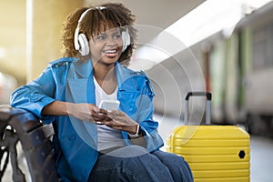 Cheerful Black Female Listening Music On Smartphone While Sitting At Railway Station