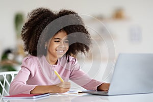 Cheerful black girl schooler doing homework, using laptop