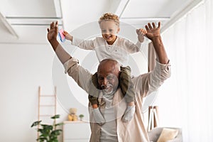 Cheerful Black Grandpa Carrying Little Grandson On Shoulders Playing Indoor