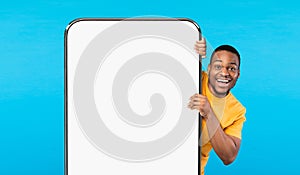 Cheerful Black Guy Peeking Out Behind Big Smartphone With White Blank Screen