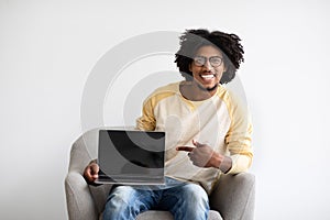 Cheerful Black Guy Pointing At Blank Laptop Screen While Sitting In Chair