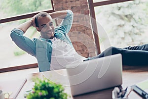 Cheerful black guy is watching at his laptop screen, at his work place, with arms behind the head, resting, smiling, in the office