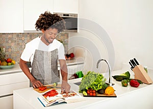 Cheerful Black Man Reading Cooking Book With Recipes At Kitchen