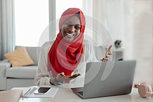 Cheerful black muslim woman in hijab having video chat with friends or online meeting, talking to laptop webcamera