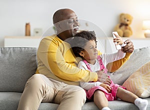 Cheerful black small girl and old man hugging and taking selfie on smartphone on sofa in living room
