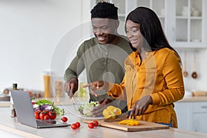 Cheerful Black Spouses Cooking Lunch In Kitchen And Checking Recipes On Laptop